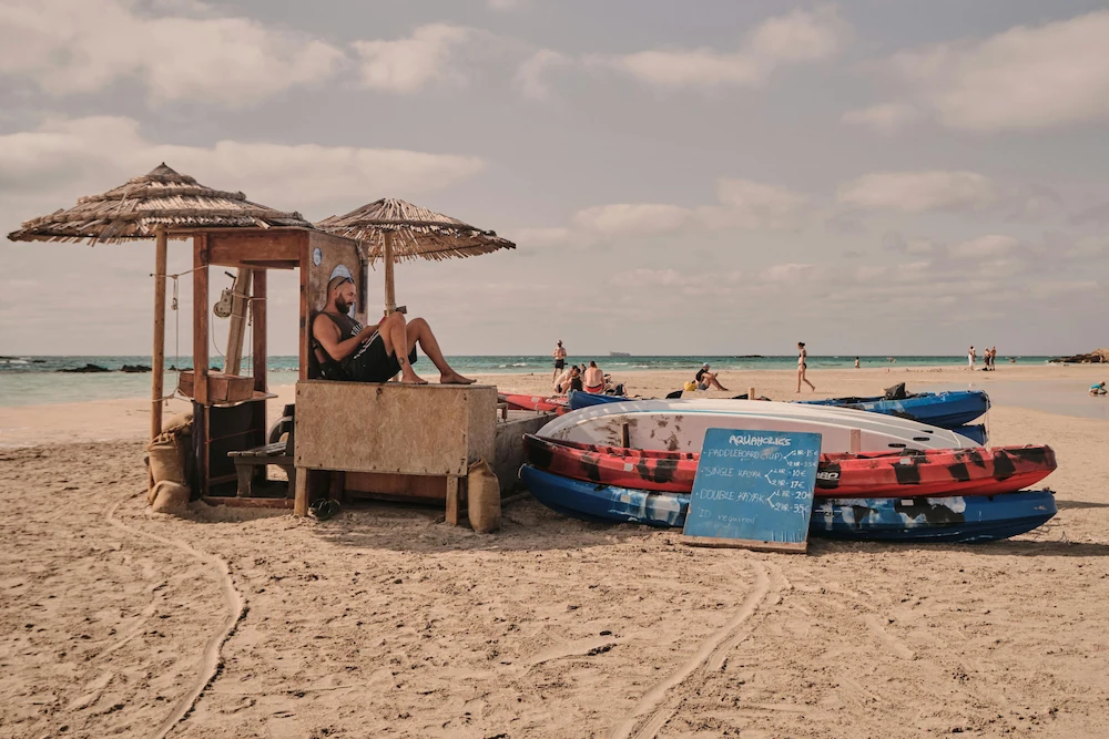 A man from our book club reading a book on a sunny beach in Crete, with colorful kayaks nearby.